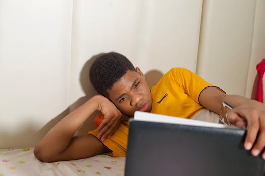 A teenage boy in a yellow shirt lying on a bed and reading indoors.