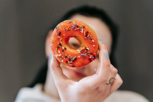 Faceless female showing tasty donut with sprinkles and looking at camera through its hole in studio