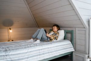Boy lying on bed in attic room, using tablet for entertainment.